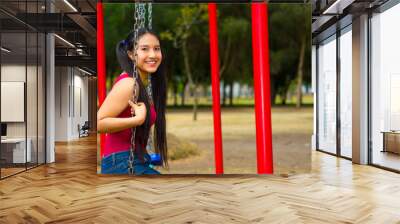 Young pretty teenage girl with pig tails wearing jeans and purple top, sitting on swing at outdoors playground, smiling happily for camera Wall mural