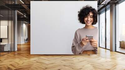 Photo of cheerful delighted woman types sms on modern cell phone device, enjoys good internet connection, using social media, isolated on grey studio wall, smiling, with copy space Wall mural