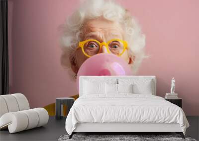 Elderly Caucasian woman in yellow glasses blowing a pink chewing gum bubble against a solid pink background with copy space Wall mural