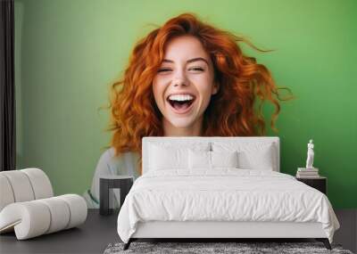 A young beautiful girl with red curly hair and a snow-white smile laughs in green T-shirt on bright green plain background. Human emotions, joy, laughter, happiness Wall mural