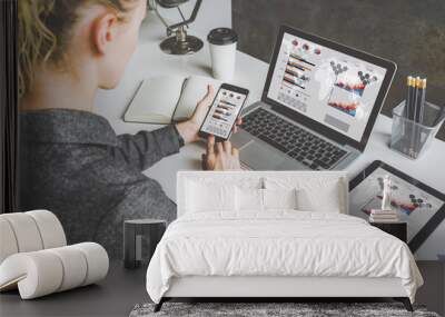 Back view. Young business woman sitting in office at table and using smartphone. On desk is laptop and tablet computer, on screen charts and graphs. Woman analyzing data. Student learning online. Wall mural