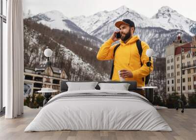 Bearded man in yellow hoodie with backpack stands on background of high snowy mountains, talking on mobile phone, drinking coffee. Wall mural