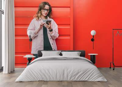 Businesswoman in glasses stands outside on red metal wall background,looking at screen of phone,uses smartphone. Wall mural