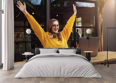 Girl rejoices in winning having raised her hands up sitting in front of laptop.Woman watches sports match on computer. Wall mural