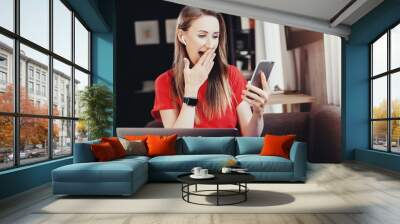 Girl sitting in front of laptop watching shocking video on smartphone. Young lady wearing wireless earphones looking at screen of mobile phone with emotions. Funny pictures, interesting posts in media Wall mural