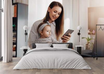 Little girl sitting on mother's knees and looking at screen of mom's smartphone. Young woman and daughter making video call in messenger app on mobile phone. Girl texting messages and comments in blog Wall mural