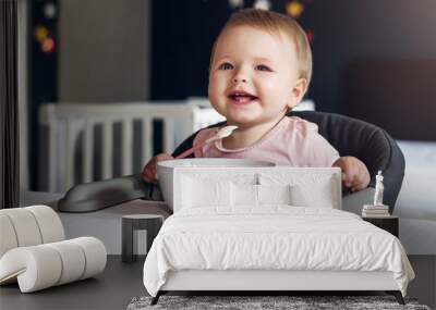 Nine-month-old smiling baby girl sits at white table in highchair and eats herself with spoon from bowl. Blurred background. Wall mural