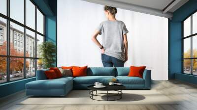 Rear view of young woman, dressed in light gray T-shirt and blue jeans, standing for light background with his hand in back pocket of jeans. Mock up. Space for text, advertising. Wall mural