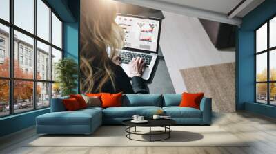 Rear view. Young businesswoman is sitting at table, working on laptop with graphs, charts, diagrams, schedules on screen Wall mural