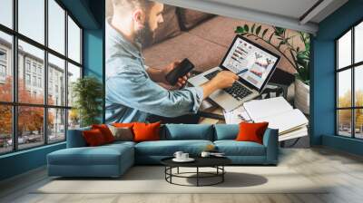 Rear view. Young hipster man, entrepreneur sits at home on couch at coffee table, holding smartphone, showing pencil on screen of laptop with graphs, charts, diagrams. Online marketing, e-learning. Wall mural