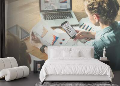 Rear view.Two young business women sitting at table.First woman signs documents,second holds graph and using smartphone.On table is laptop and tablet computer. Diagrams on screen of laptop and phone. Wall mural