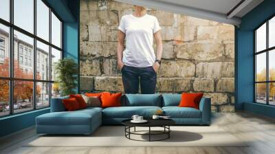 Summer day. Front view. Young woman in a white T-shirt and blue pants standing on a background of a stone wall with his hands in his pockets. Cropped image. Mock up. Wall mural