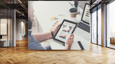 Tablet computer with graphs, diagrams and charts on screen in hands of young bearded businessman sitting at table Wall mural