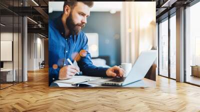 Young bearded businessman works on computer, takes notes in notebook, analyzes information, makes business plan. Freelancer, entrepreneur works at home. Online marketing, education for adults. Wall mural