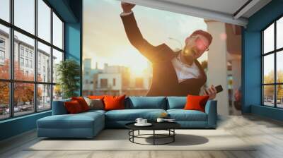 Attractive satisfied young man clenching fist jumping with happiness at sunset. Outdoor portrait happy businessman i proud of victory smiling and having fun in the street. Wall mural