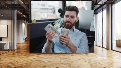 Cheerful smiling young man business manager counting bundle of money dollars celebrating profits financial success in modern office company. Wall mural