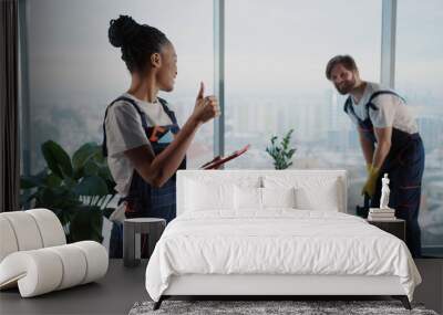 Cleaning services team. Young man using vacuum cleaner tidying office workspace. Female african american cleaning employee using tablet computer. Wall mural