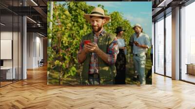 Handsome happy farmer in hat using application on smartphone in behind young luxury people tasting grapes and wine on vineyard. Wall mural