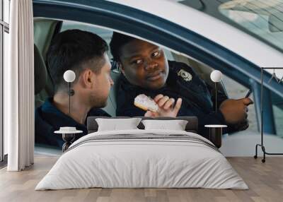 Two police officers best friends talking heart to heart sitting inside patrol car during a break at work. Handsome policeman eating doughnuts and his colleague using a smartphone. Wall mural