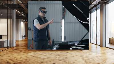 A male mechanic in a virtual reality headset gesturing and standing near the open hood of a car, while making a diagnostics in a car service Wall mural