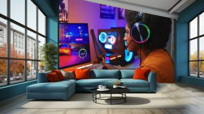 African American guy, teenage gamer in headphones plays in online 3D race game on modern powerful PC in neon cozy room. Video game live streaming or professional eSports tournament. Gaming at home. Wall mural