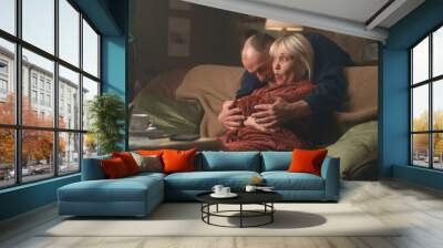 Aged husband embracing and kissing surprised wife in bathrobe resting on sofa in living room in evening at home Wall mural