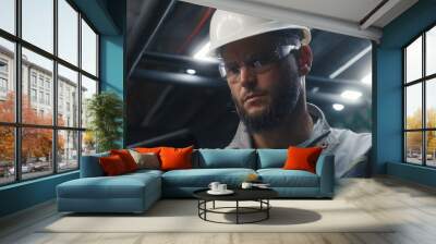 Close up of professional Caucasian engineer using digital tablet computer on industrial manufacturing factory. Heavy industry specialist in safety uniform, goggles and hard hat works on modern plant. Wall mural