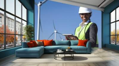 Female engineer wearing hard hat standing with digital tablet against wind turbine on sunny day and looking at camera smilingly Wall mural