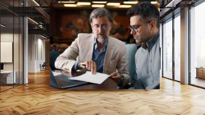 Mature man using laptop, talking with serious businessman, showing papers, documents. Business meeting in office restaurant. Signing contract for new economic strategy of company Wall mural