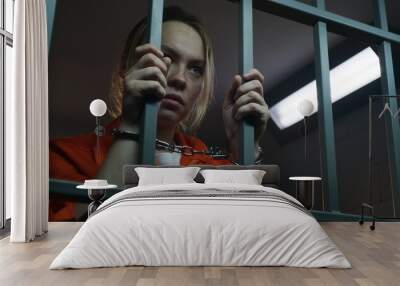 Shocked female prisoner in orange uniform holds metal bars, stands in prison cell in handcuffs. Woman criminal serves imprisonment term in jail. Depressed inmate in correctional facility. Portrait. Wall mural