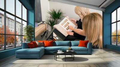 Two women are sitting in chairs looking at a book with hair color charts Wall mural