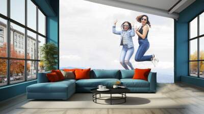 two attractive latin american young women outdoors with sunglasses smiling happy and jumping high looking to camera in a summer day vacation trip with nature in background Wall mural