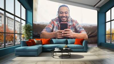 Smiling young latino man in his thirties sitting on his couch at home using his smart phone. Wall mural