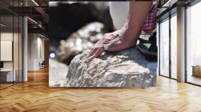 A silver ring with a cross and diamonds on the finger of an elder man who sits on a coastal rock. Wall mural