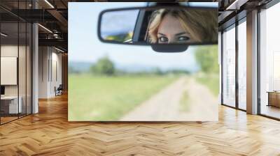 The beautiful eyes of the young driver woman are reflected in the rearview mirror. Blurred road and landscape is in the background. Wall mural