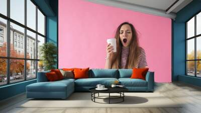 Cute girl with a glass of coffee in hand is yawning. She can't wake up. Wall mural