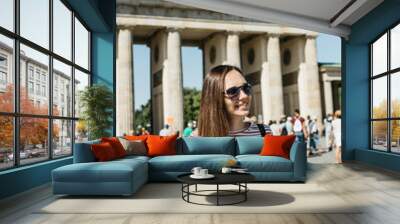 Portrait of a young beautiful positive smiling stylish tourist girl in Berlin in Germany. Brandenburg Gate and unrecognizable blurred people in the background. Wall mural