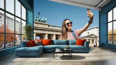 Young beautiful positive girl makes selfie against the background of the Brandenburg Gate in Berlin in Germany or takes pictures of sights. Wall mural