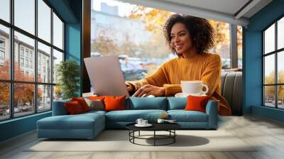 Smiling woman working on laptop in cozy cafe with coffee, daytime, casual autumn attire, productivity and relaxation concept Wall mural