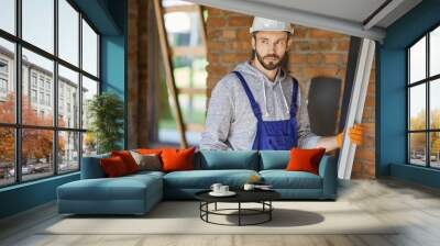 Attractive male builder wearing overalls and hard hat looking away while holding a metal stud for drywall on interior site building Wall mural