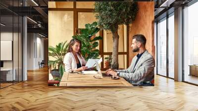 Business lunch. Man sitting at table working on laptop at restaurant while woman reading documents smiling cheerful Wall mural