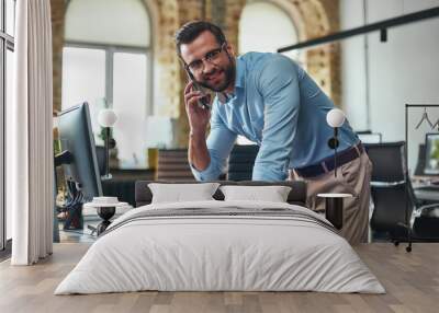 Business talk. Portrait of young and cheerful bearded man in eyeglasses talking on smart phone and using computer while standing in the modern office Wall mural