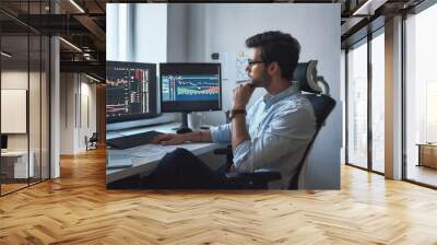 Busy working day. Side view of successful trader or businessman in formal wear and eyeglasses working with charts and market reports on computer screens in his modern office Wall mural