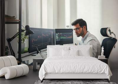 Busy working day. Young bearded trader in eyeglasses working with laptop while sitting in his modern office in front of computer screens with trading charts. Wall mural