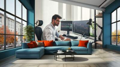 Busy working day. Young bearded trader in eyeglasses working with laptop while sitting in his modern office in front of computer screens with trading charts. Wall mural