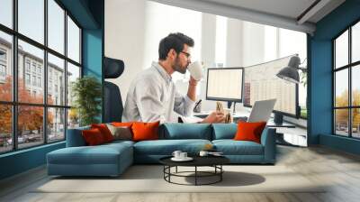 Coffee break. Young bearded businessman or trader is drinking a coffee and eating a sandwich while looking at monitor screen with financial data in his modern office Wall mural