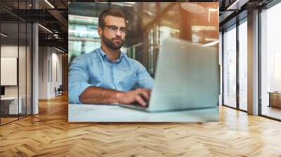 Comfortable working place. Young bearded businessman in eyeglasses and formal wear using laptop while sitting in modern office Wall mural