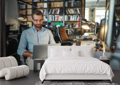 Comfortable workplace. Portrait of young and successful bearded man in eyeglasses working with laptop while sitting in modern office Wall mural