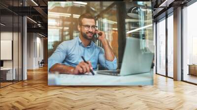 Customer support. Portrait of young and cheerful businessman in eyeglasses and formal wear talking on the phone and smiling while sitting in the office Wall mural