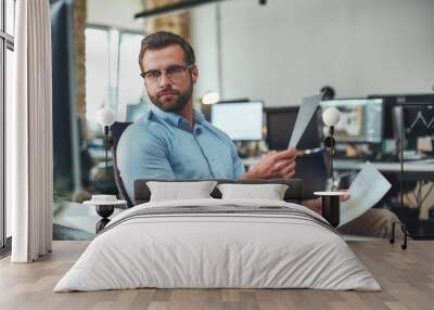 Examining the reports. Busy and focused bearded man in eyeglasses and formal wear holding documents and looking at computer monitor while sitting in the modern office Wall mural